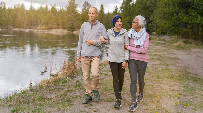 An older couple link arms with a younger woman wearing a head scarf as they walk next to a pond 