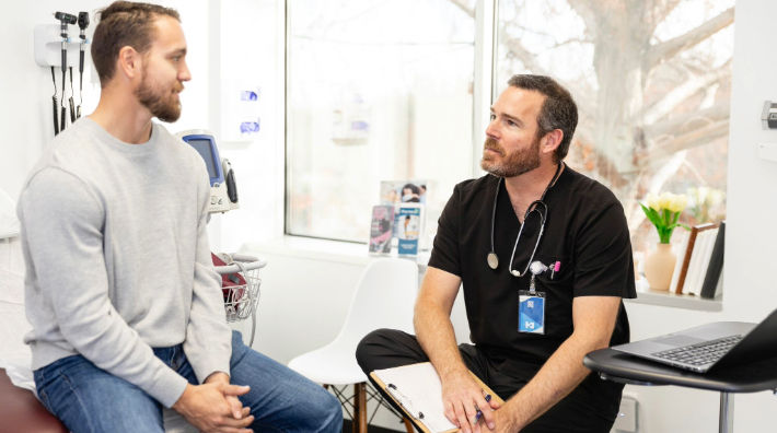 A bearded man in a gray sweatshirt sits on an exam table facing a male doctor wearing black scrubs in a doctor’s office.