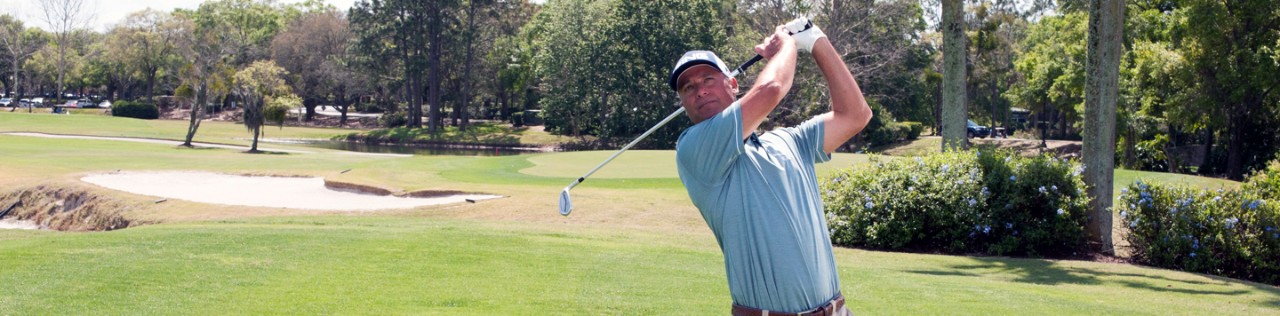 A man in a blue shirt playing golf, swinging a club on a lush green golf course